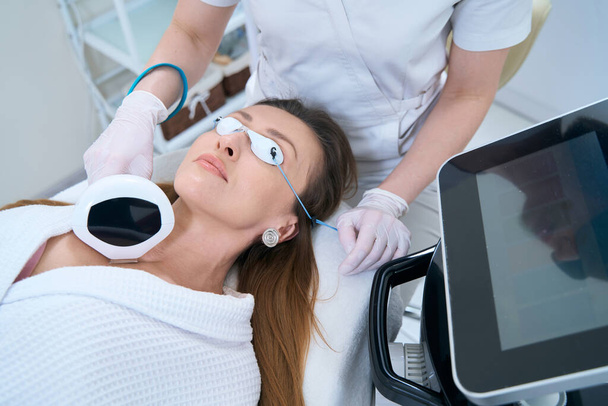 Жінка в захисних окулярах лежить на процедурі фотоомолодження шкіри шиї
 - Фото, зображення