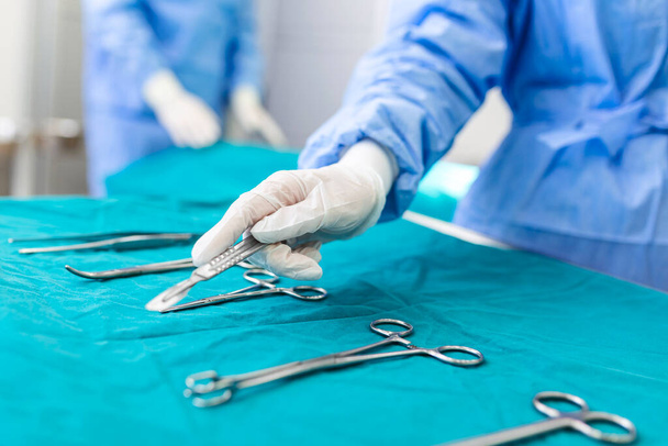 Nurse hand taking surgical instrument for group of surgeons at background operating patient in surgical theatre. Steel medical instruments ready to be used. Surgery and emergency concept - Photo, Image