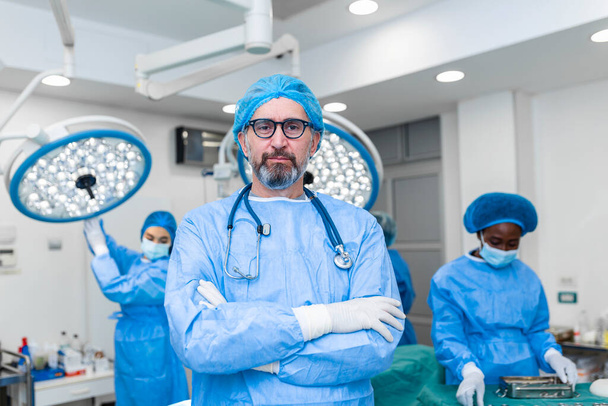 Portrait of male surgeon in operation theater looking at camera. Doctor in scrubs and medical mask in modern hospital operating theater. - 写真・画像