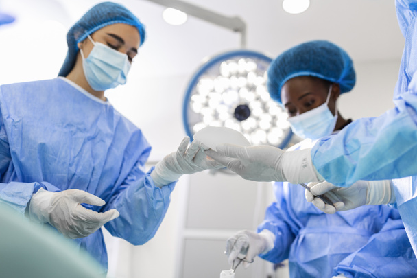 Surgeon and his assistant performing cosmetic surgery in hospital operating room. Surgeon in mask during medical procadure. Breast augmentation, enlargement, enhancement - Foto, Bild