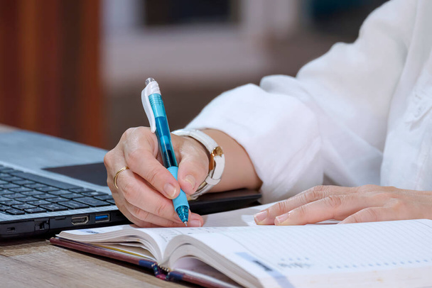 Fechar-se da mão de mulher de negócios que escreve no caderno com o computador portátil na mesa de escritório - Foto, Imagem