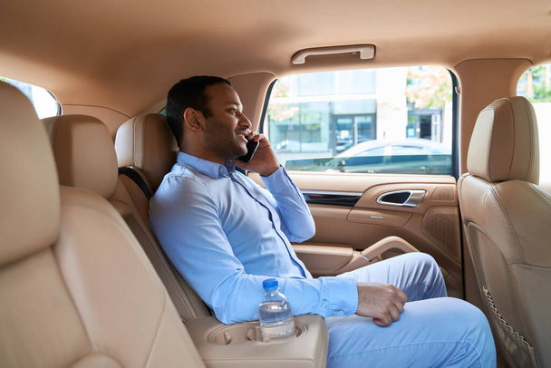 Joyous young man seated in backseat of auto talking on his smartphone - Foto, Imagen