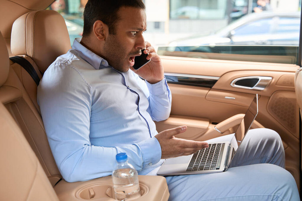 Shocked young man seated in backseat of auto staring at his computer during phone call - Photo, Image