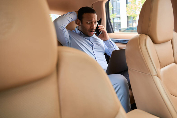Businessperson seated in backseat of taxicab goggling at his computer display during phone call - Photo, Image