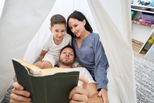 Dad, mom and son comfortably settled on the pillows, father is reading, mother and boy are listening enthusiastically - Foto, Bild