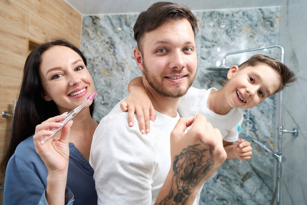 Family of three cheerfully brushes their teeth in bathroom, mom, dad and son have a good mood, they have fun together - Photo, Image