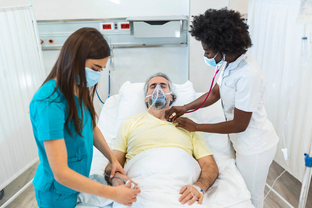 Jovem médico e enfermeiro vestindo uma máscara cirúrgica verificando um paciente sênior do sexo masculino usando uma máscara de oxigênio de pressão positiva para ajudar a respirar em uma cama de hospital durante a pandemia de Covid-19 - Foto, Imagem