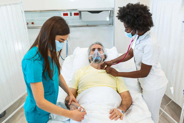 Médicos com máscaras protetoras estão examinando o paciente idoso infectado no hospital. No Hospital Senior Paciente Descansa com máscara de oxigênio, deitado na cama. - Foto, Imagem