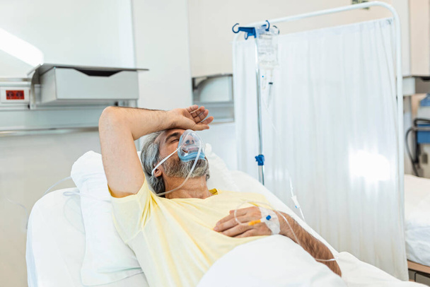 Pacjent z koronawirusem odwraca wzrok podczas odpoczynku na szpitalnym łóżku. Facet nosi maskę tlenową. Jest w szpitalu podczas pandemii.. - Zdjęcie, obraz