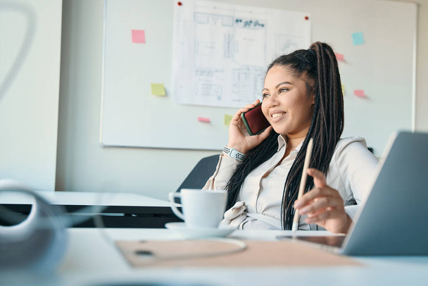 Smiling female entrepreneur sitting at office desk and looking into distance during phone talk - Photo, Image