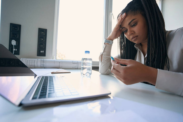 Tired female entrepreneur sitting at office desk and looking at drug capsule in her hand - Foto, Imagem