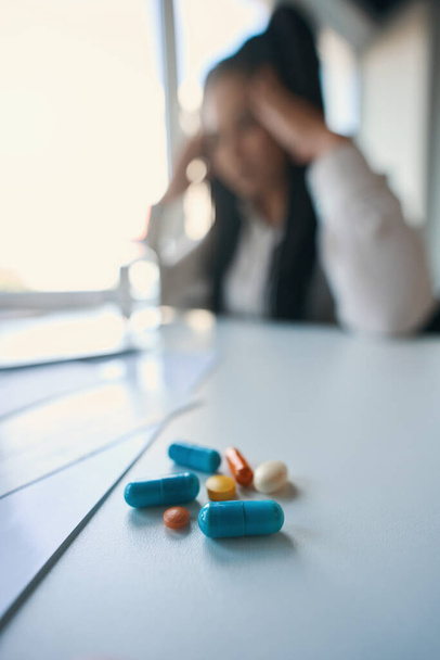 Defocused photo of company employee seated at table with pile of pharmaceutical drugs touching temples with hands - Fotoğraf, Görsel