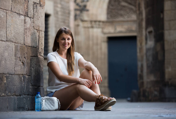 Happy young woman strolling around city sitting near old stone wall - Zdjęcie, obraz