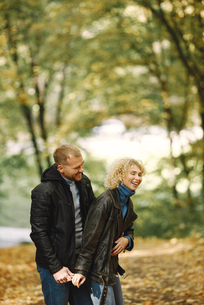 Blonde curly woman and man standing in autumn forest and hugging. Couple wearing leather jackets and blue sweaters. Photo of romantic couple in a forest. - Fotoğraf, Görsel