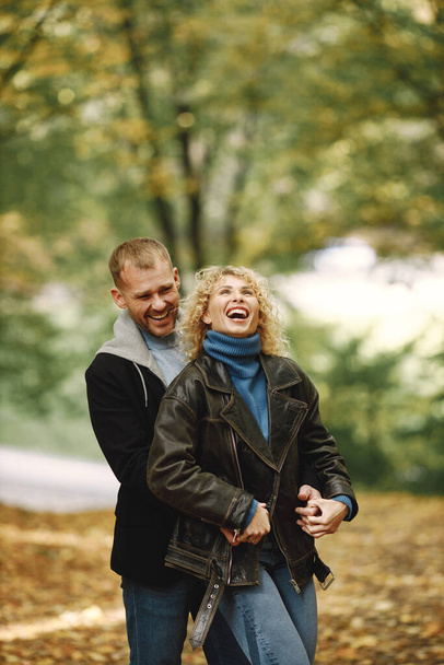 Blonde curly woman and man standing in autumn forest and hugging. Couple wearing leather jackets and blue sweaters. Photo of romantic couple in a forest. - Foto, Imagen