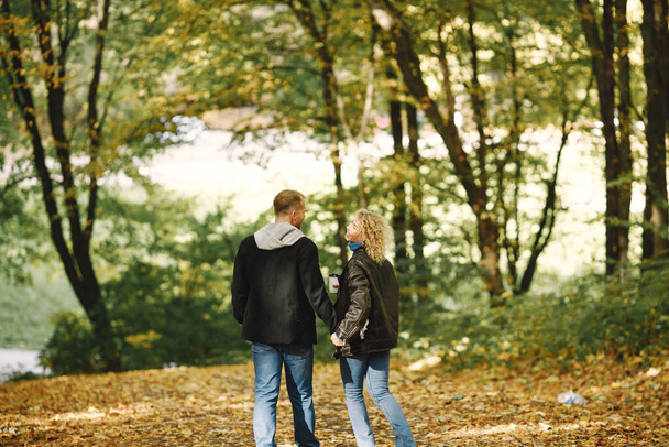 Blonde curly woman and man holding hands in autumn forest and hugging. Couple wearing leather jackets and blue sweaters. Photo of romantic couple in a forest. - Foto, Bild