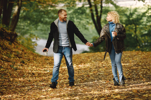 Blonde curly woman and man holding hands in autumn forest and hugging. Couple wearing leather jackets and blue sweaters. Photo of romantic couple in a forest. - Foto, Imagen