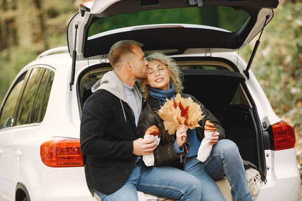 Blonde curly woman and man sitting in a trunk in car in autumn forest and hugging. Couple wearing leather jackets and blue sweaters. Photo of romantic couple in a forest eating a croissants. - Fotoğraf, Görsel