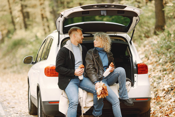 Blonde curly woman and man sitting in a trunk in car in autumn forest and hugging. Couple wearing leather jackets and blue sweaters. Photo of romantic couple in a forest eating a croissants. - Photo, Image