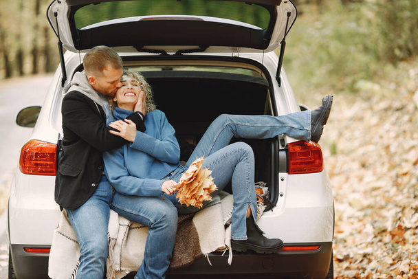 Blonde curly woman and man sitting in a trunk in car in autumn forest and hugging. Man wearing black leather jacket and woman blue sweater. Photo of romantic couple in a forest. - Фото, зображення