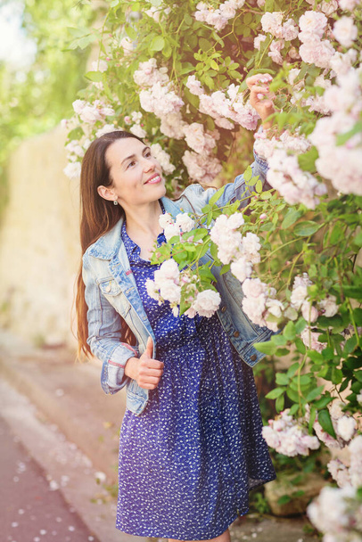 Lifestyle portrait of young stylish woman staying on the street in old town and touching the flowers of white roses, wearing blue dress and denim jacket - Φωτογραφία, εικόνα