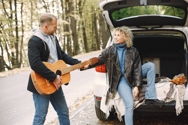 Blonde curly woman and man sitting in a trunk in car in autumn forest with a guitar. Couple wearing leather jackets and blue sweaters. Photo of romantic couple in a forest. - Zdjęcie, obraz