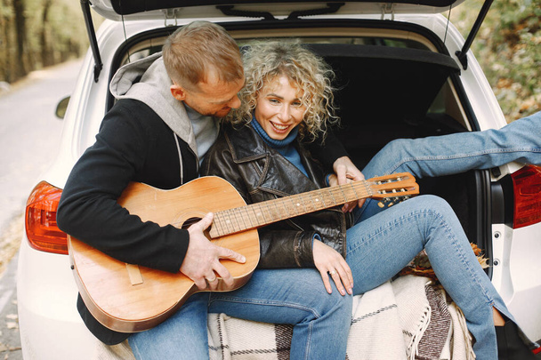 Blonde curly woman and man sitting in a trunk in car in autumn forest with a guitar. Couple wearing leather jackets and blue sweaters. Photo of romantic couple in a forest. - Фото, изображение