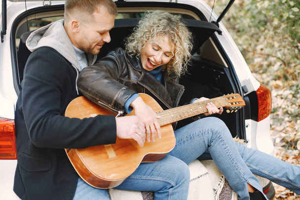 Blondynka kręcone kobieta i mężczyzna siedzi w bagażniku w samochodzie w jesiennym lesie z gitarą. Para w skórzanych kurtkach i niebieskich swetrach. Zdjęcie romantycznej pary w lesie. - Zdjęcie, obraz