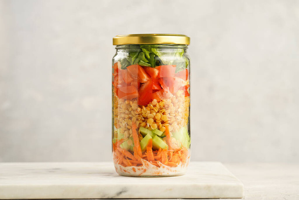 Trendy Fresh detox summer salad layered in a glass mason jar: yoghurt dressing, carrots, cucumber, yellow lentils, red bell pepper, green leaves and herbs, long silver spoon, on white background - Fotoğraf, Görsel