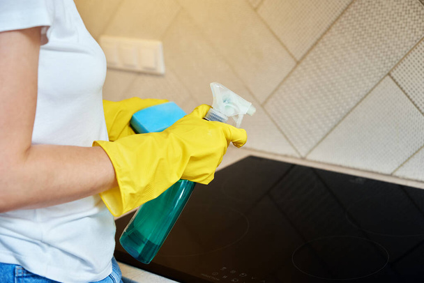 Schoonmaken inductiekachel. Vrouw in geel rubber handschoenen reinigt keuken inductie kookplaat met schoonmaken sparay en spons. Duidelijke keukenapparatuur - Foto, afbeelding