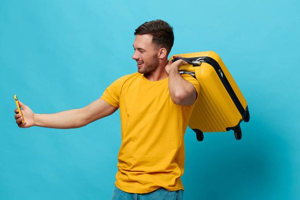 Happy cheerful tanned handsome man in yellow t-shirt doing selfie video call with phone hold suitcase posing isolated on blue studio background. Copy space Banner Mockup. Trip journeys concept - Photo, Image