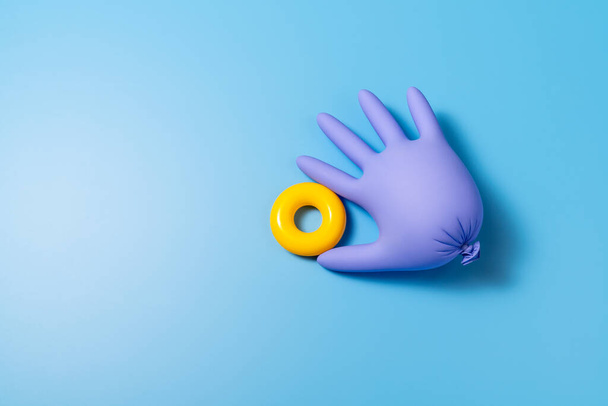 Violet inflated latex glove with a plastic yellow ring between its fingers lies on a vibrant blue background in a studio. Top view horizontal photo. - Photo, image