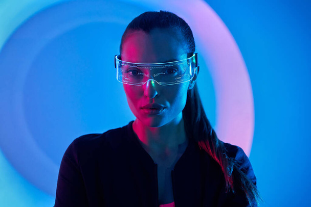 Confident young woman in futuristic glasses standing against colorful background - 写真・画像