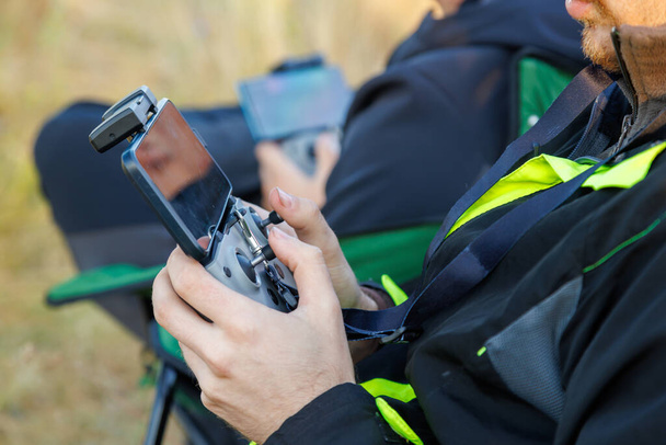 スティックや携帯電話を使って遠隔操作するドローンオペレーターの手は、ぼんやりとした野生の草の背景を昼間監視します - 写真・画像