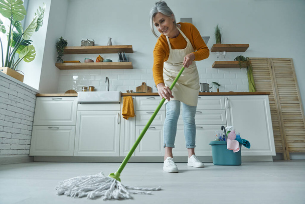 Full length of senior woman cleaning floor with mop while standing at the domestic kitchen - Φωτογραφία, εικόνα