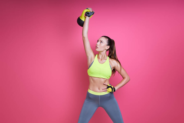 Concentrated young woman in sports clothing carrying kettlebell against pink background - Фото, изображение