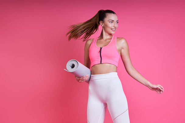 Beautiful young woman in sports clothing carrying an exercise mat against pink background - Fotoğraf, Görsel