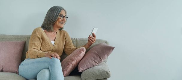 Elegant senior woman using smart phone and smiling while relaxing on the comfortable couch - Фото, изображение