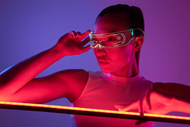 Beautiful woman in futuristic glasses leaning at the LED lamp against dark background - Fotó, kép