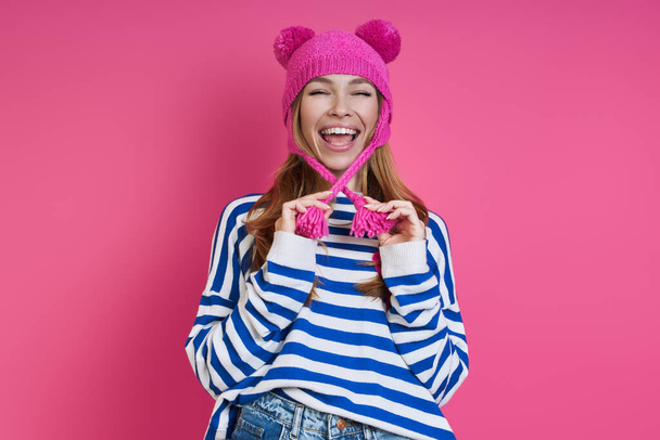 Playful young woman adjusting her pink hat and smiling while standing against colored background - Photo, Image