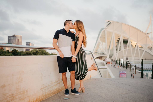 A couple of tourists are having fun with modern urban space in the background on a date at the sunset in Valencia. A girl is kissing her boyfriend in the evening in Spain - Foto, immagini