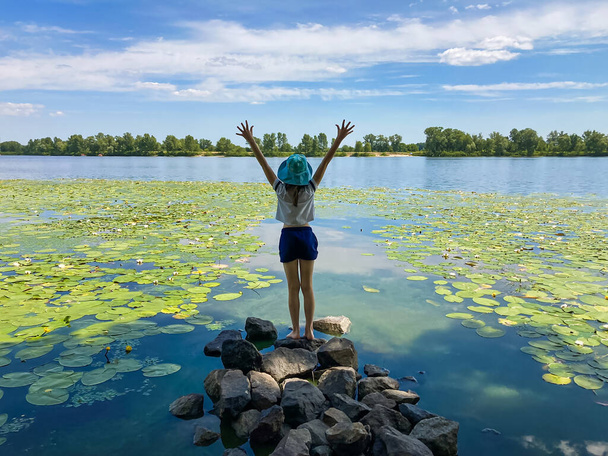 Achteraanzicht van een tienermeisje met opgeheven armen, staande op de oever van de rivier op een zomerdag. Het concept van vrede, succes en overwinning - Foto, afbeelding