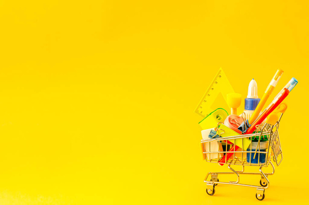 Assorted stationery in a shopping trolley on a yellow background with place for text. School preparation concept, flat lay, copy space. View from above. - Фото, изображение