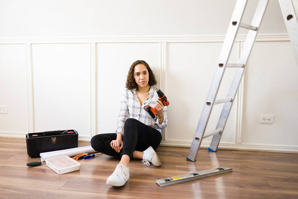 Porträt einer handlichen jungen Frau, die in die Kamera blickt, während sie mit Bohrmaschine und Leiter zu Hause Reparaturen durchführt  - Foto, Bild