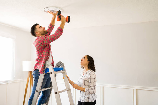 Happy young man drilling the ceiling and putting up a new lamp and lighting with his partner - Foto, Imagem