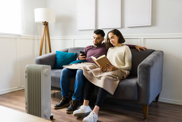 Relaxed beautiful couple resting together on the couch with a blanket and reading a book in the living room with a heater - 写真・画像
