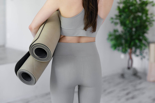 Young beautiful woman holding yoga mats while standing in a light studio. Rear view of a slim young female body. - Photo, Image