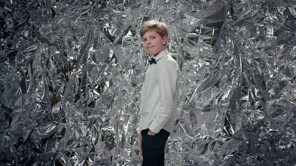 Stylish Boy In White Shirt And Black Pants. He Stands On Background Of Shiny Silver Surface. He Has Butterfly Around His Neck. Boy Holds His Hands In His Pockets. Celebration. - Foto, afbeelding