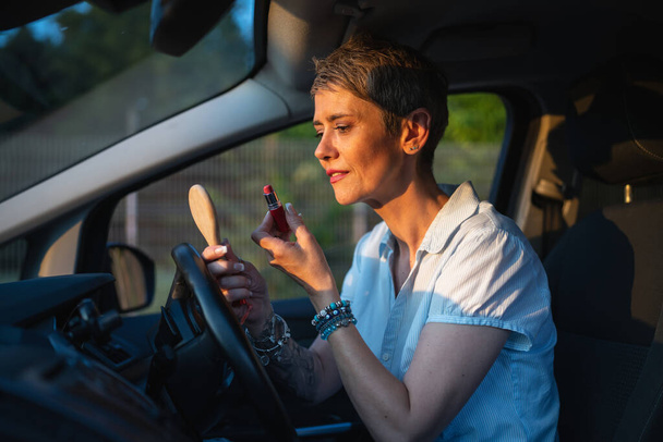 Jedna žena zralý kavkazský žena businesswoman sedí v autě uvedení rtěnky fixace opravy make-up na její tváři, zatímco čeká v létě večer skuteční lidé kopírovat prostor šedé vlasy - Fotografie, Obrázek