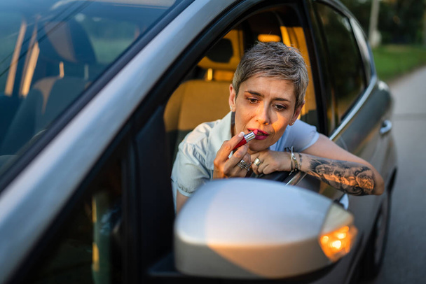 Jedna žena zralý kavkazský žena businesswoman sedí v autě uvedení rtěnky fixace opravy make-up na její tváři, zatímco čeká v létě večer skuteční lidé kopírovat prostor šedé vlasy - Fotografie, Obrázek